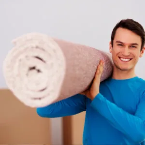 A man stands holding a large roll of carpet, showcasing its texture and color against a neutral background.