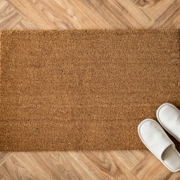 Paillasson marron en fibres naturelles posé sur un sol en bois, avec des chaussons blancs à côté.