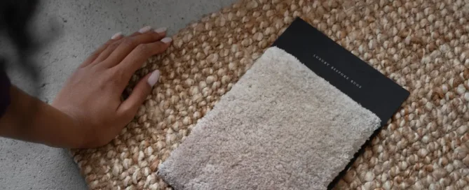 Main touchant un échantillon de tapis beige posé sur un tapis tissé en fibres naturelles.