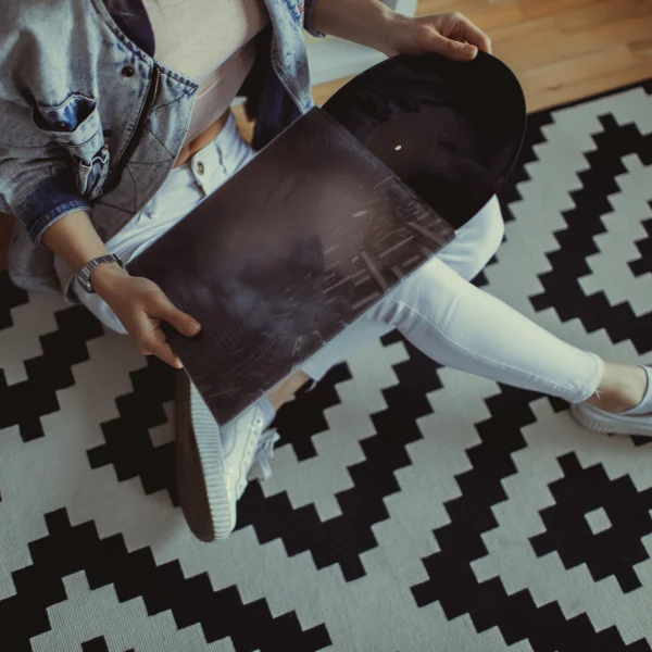 Personne assise sur un tapis géométrique noir et blanc tenant un disque vinyle, dans une ambiance lifestyle moderne.